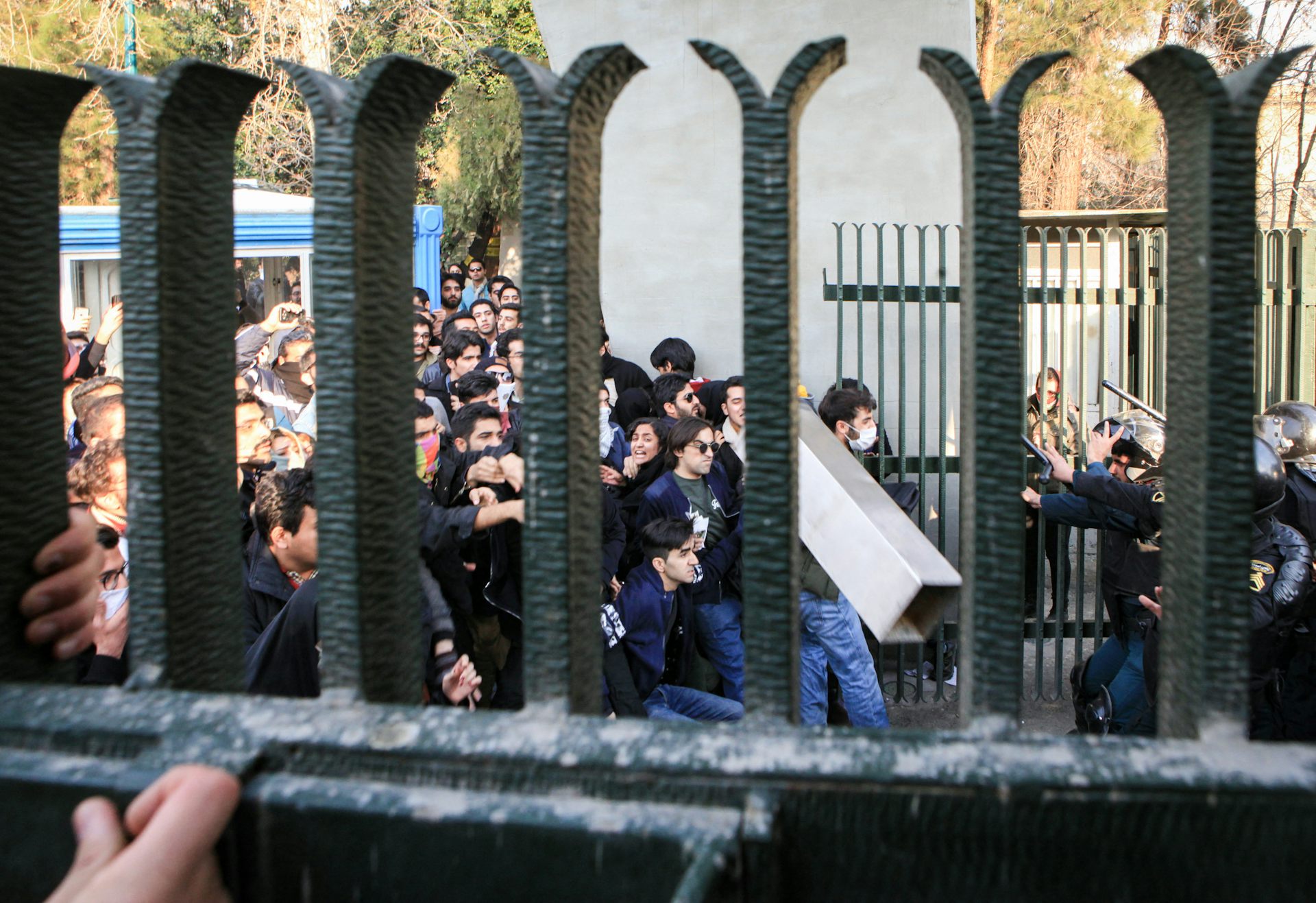 A group of young people push together against police in black, as seen through the spaces of a large fence.