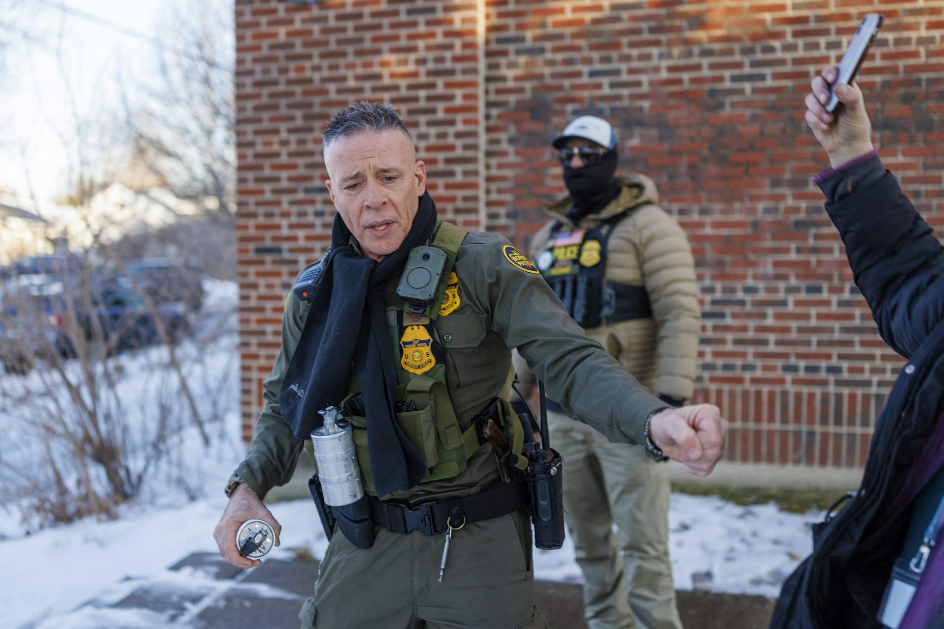 A man wearing a green vest and pants stands near another man also in a green uniform, in front of a red brick building.