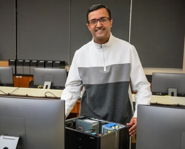 Sumeet Dave, a South Asian man with short black hair, wearing glasses and smiling in a computer lab