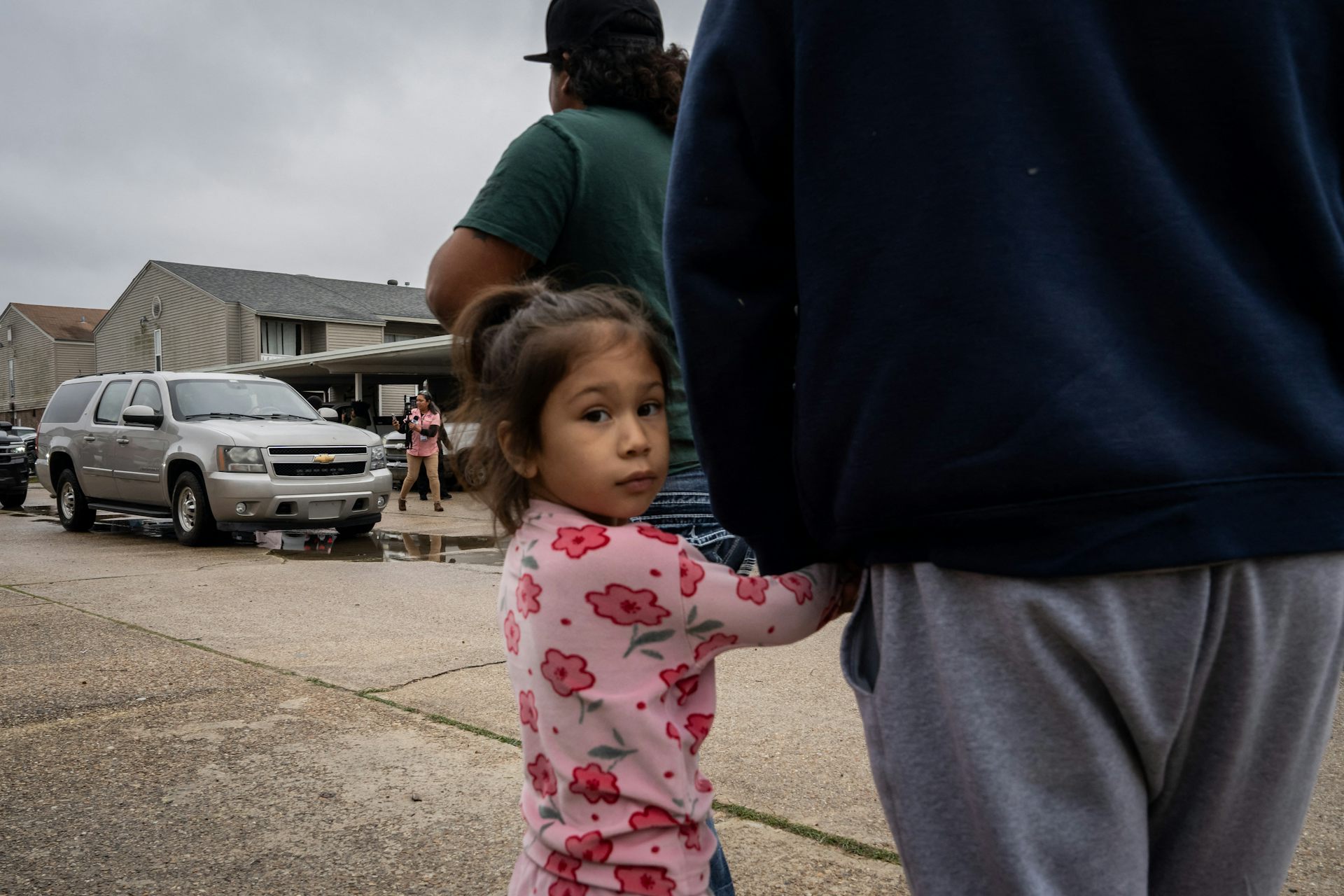 A young girl wearing a pink shirt holds an adult's hand and looks directly at the camera. She stands on a street near a parked gray SUV.