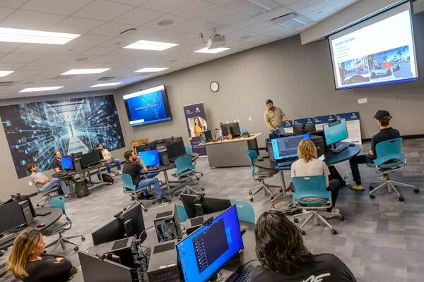 Students sit in turquoise chairs in front of computers. A professor stands behind a desk at the front of the room in front of a projector. 