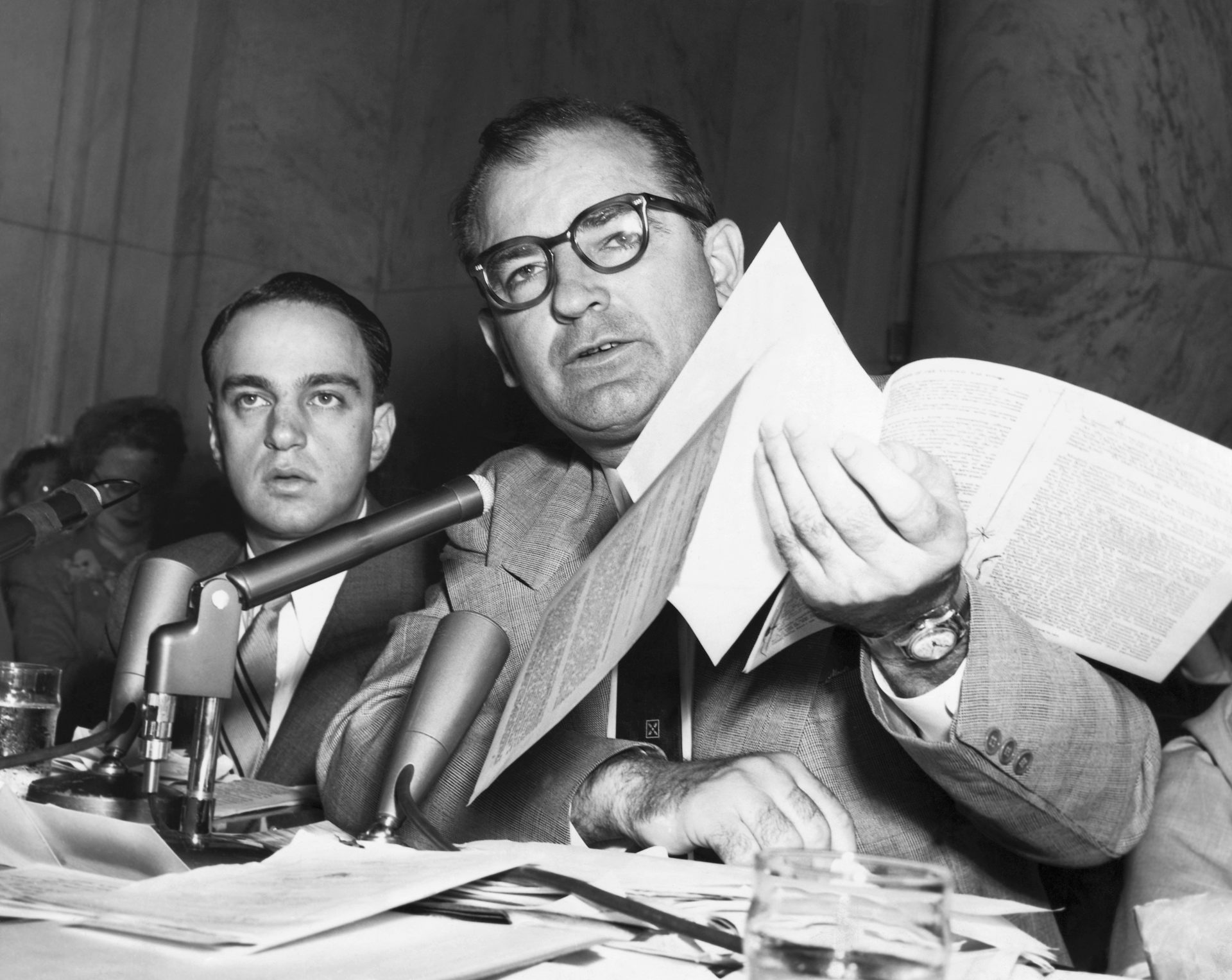 A man in a black-and-white photo wears glasses and holds up papers toward a microphone. He sits next to another man.