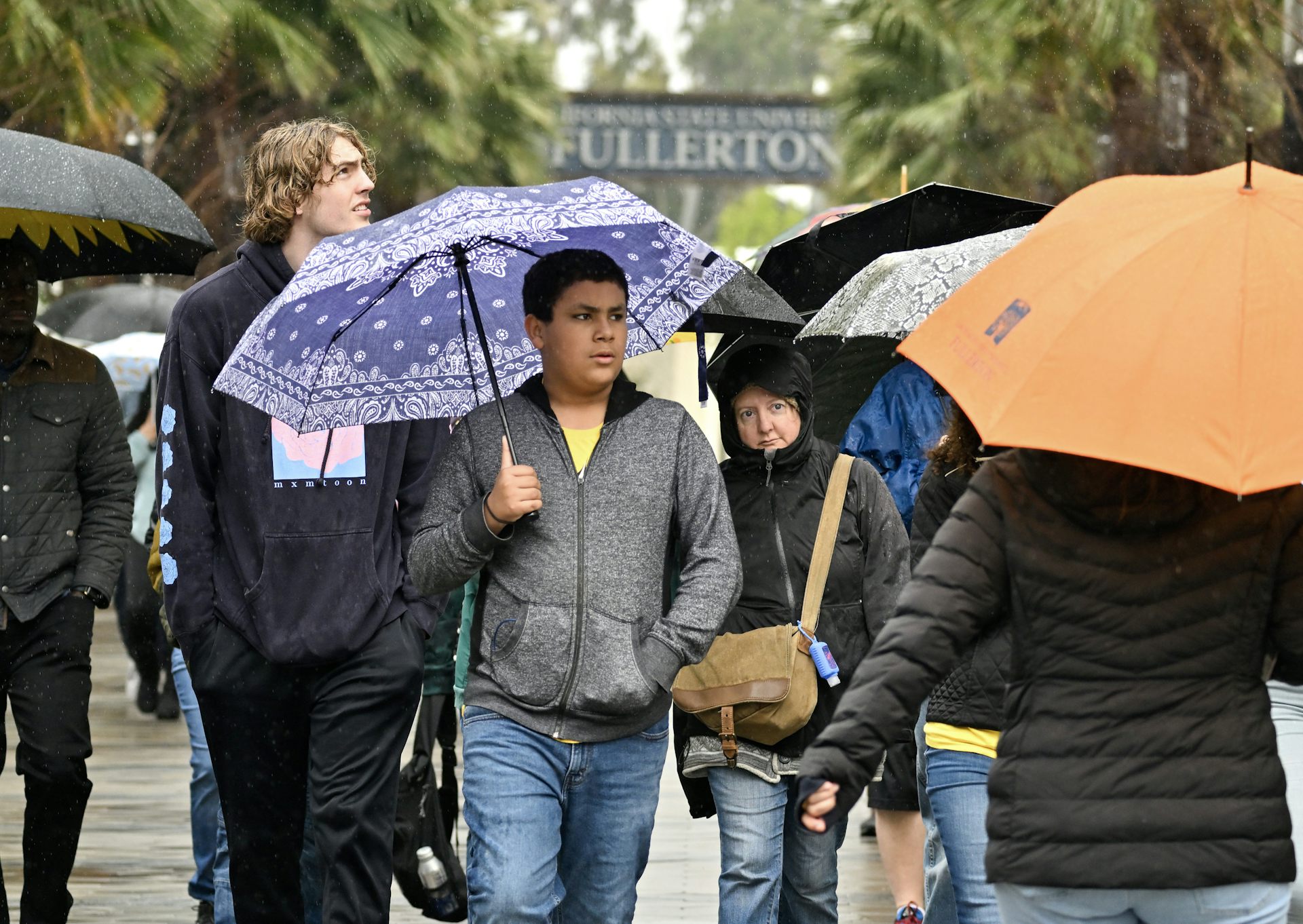 People hold umbrellas and walk together in the rain near palm trees, in front of a large sign that days Fullerton