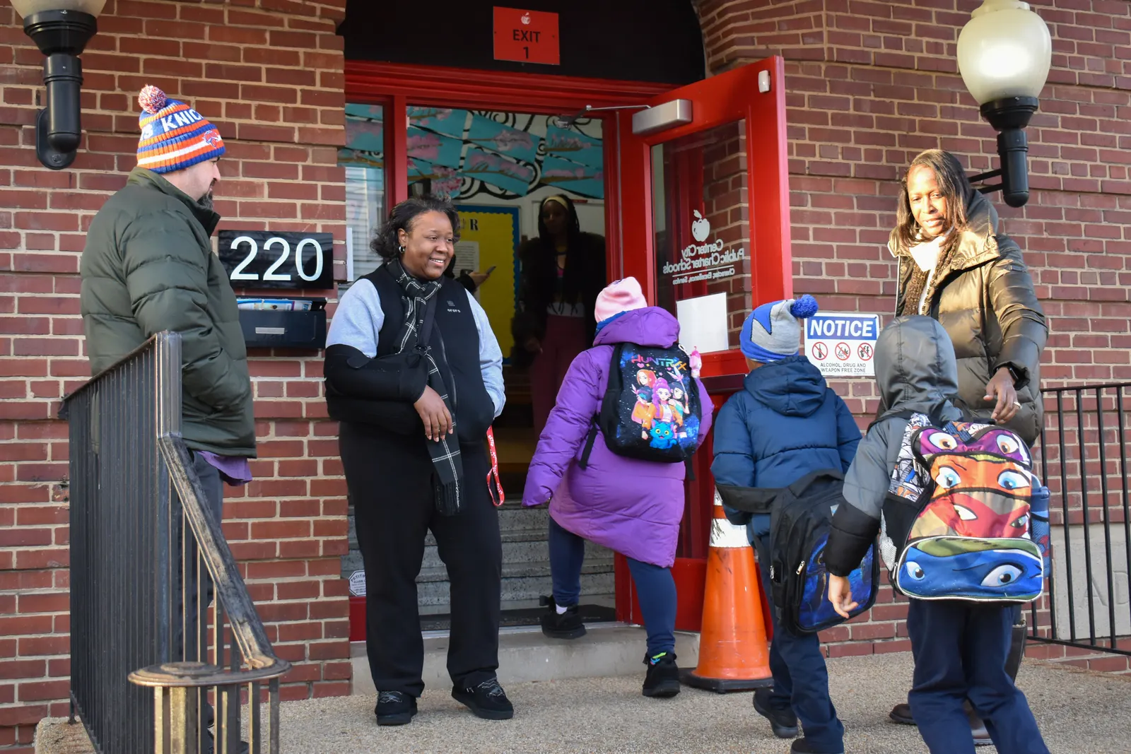 Three adults are standing at the entrance of a school greeting students as they walk into the school.