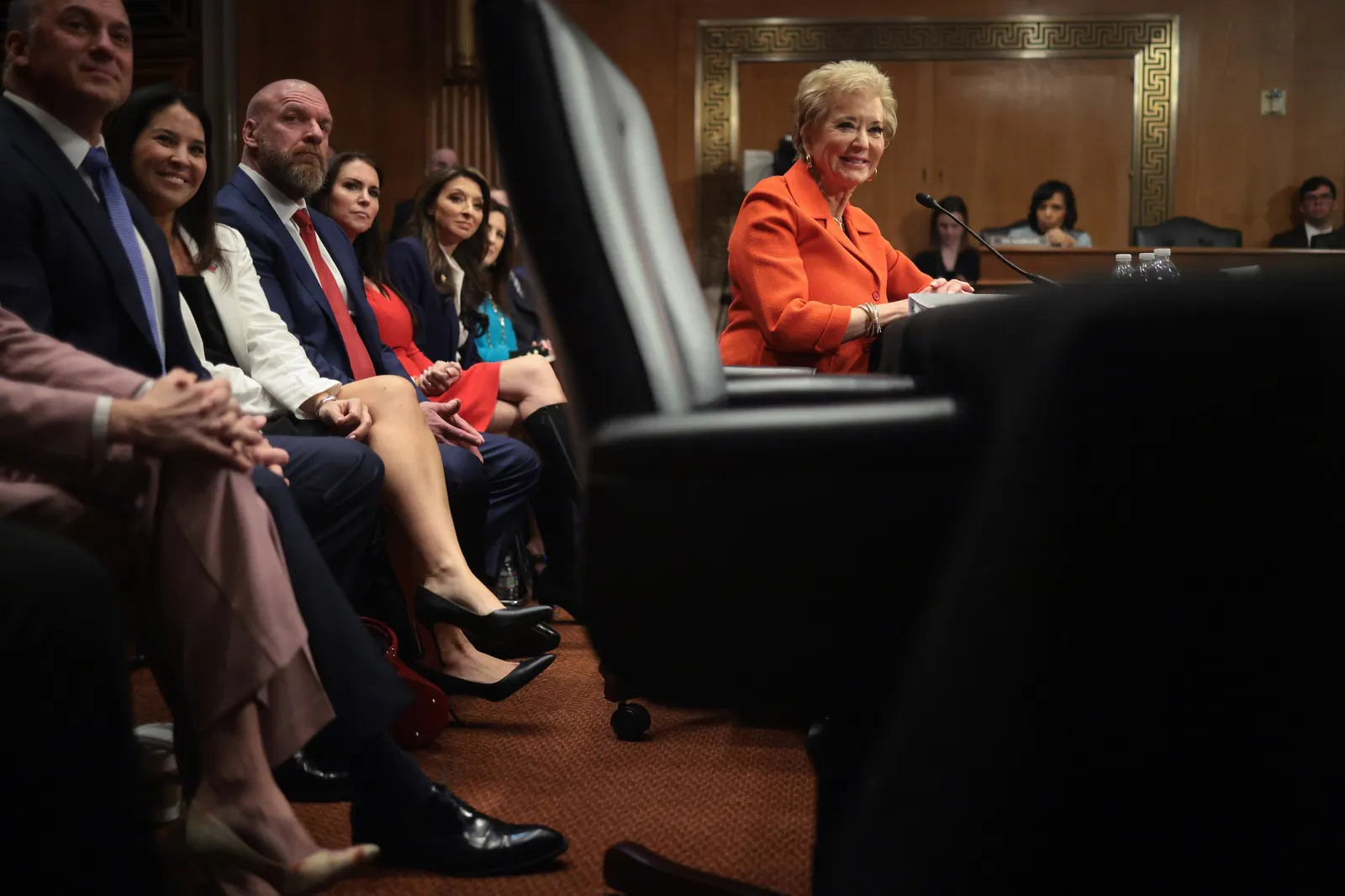 A person sits at a table with a microphone in a room with wood paneling. A row of people are seated behind the person.