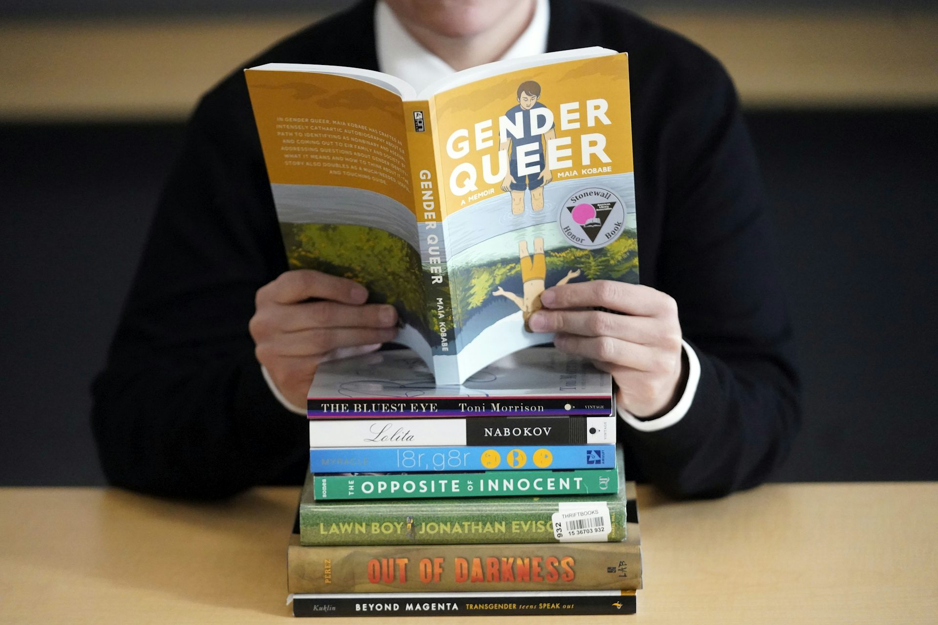 Person reading the book 'Genderqueer' atop a stack of other challenged books