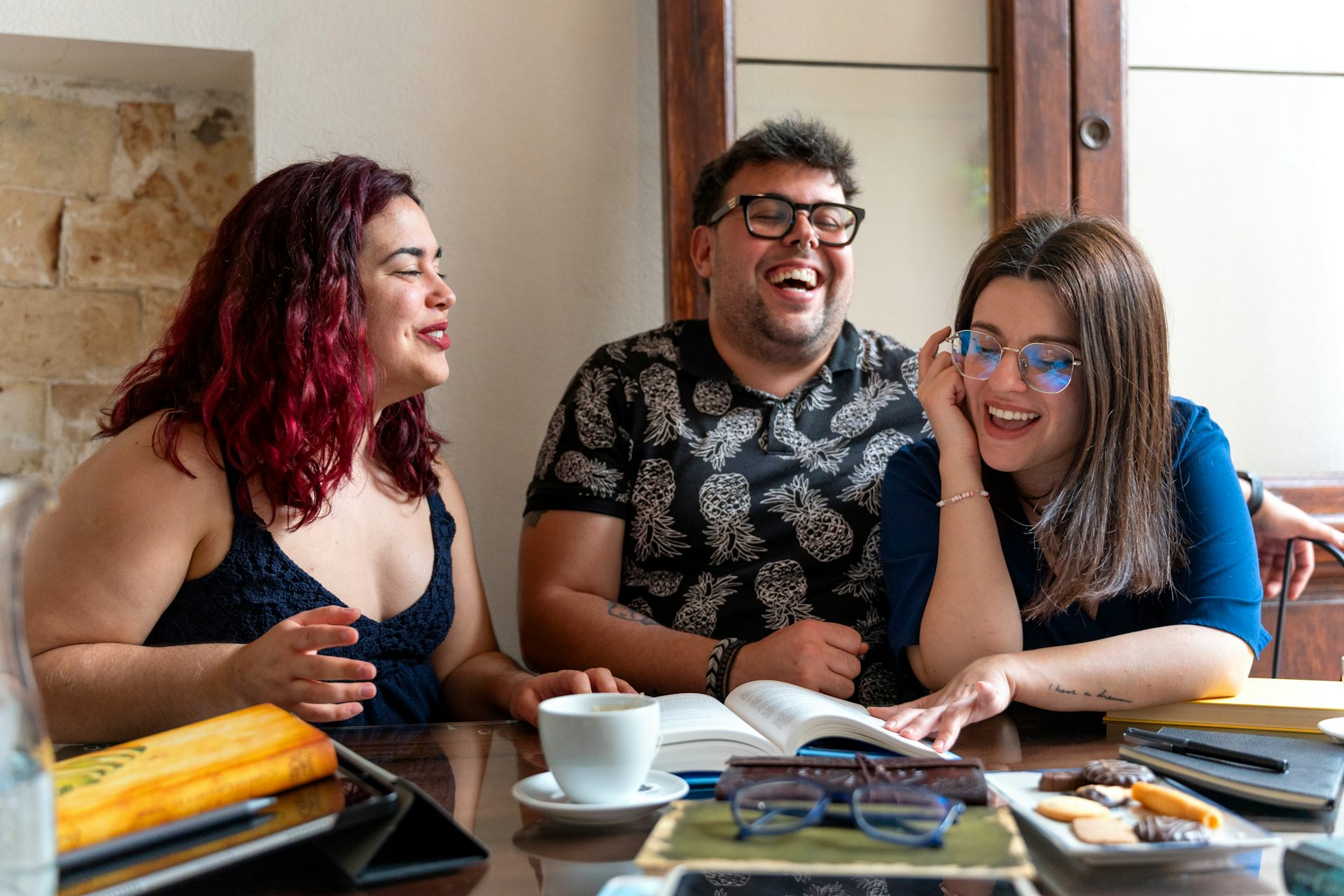 three people laughing together at a table, with books open in front of them
