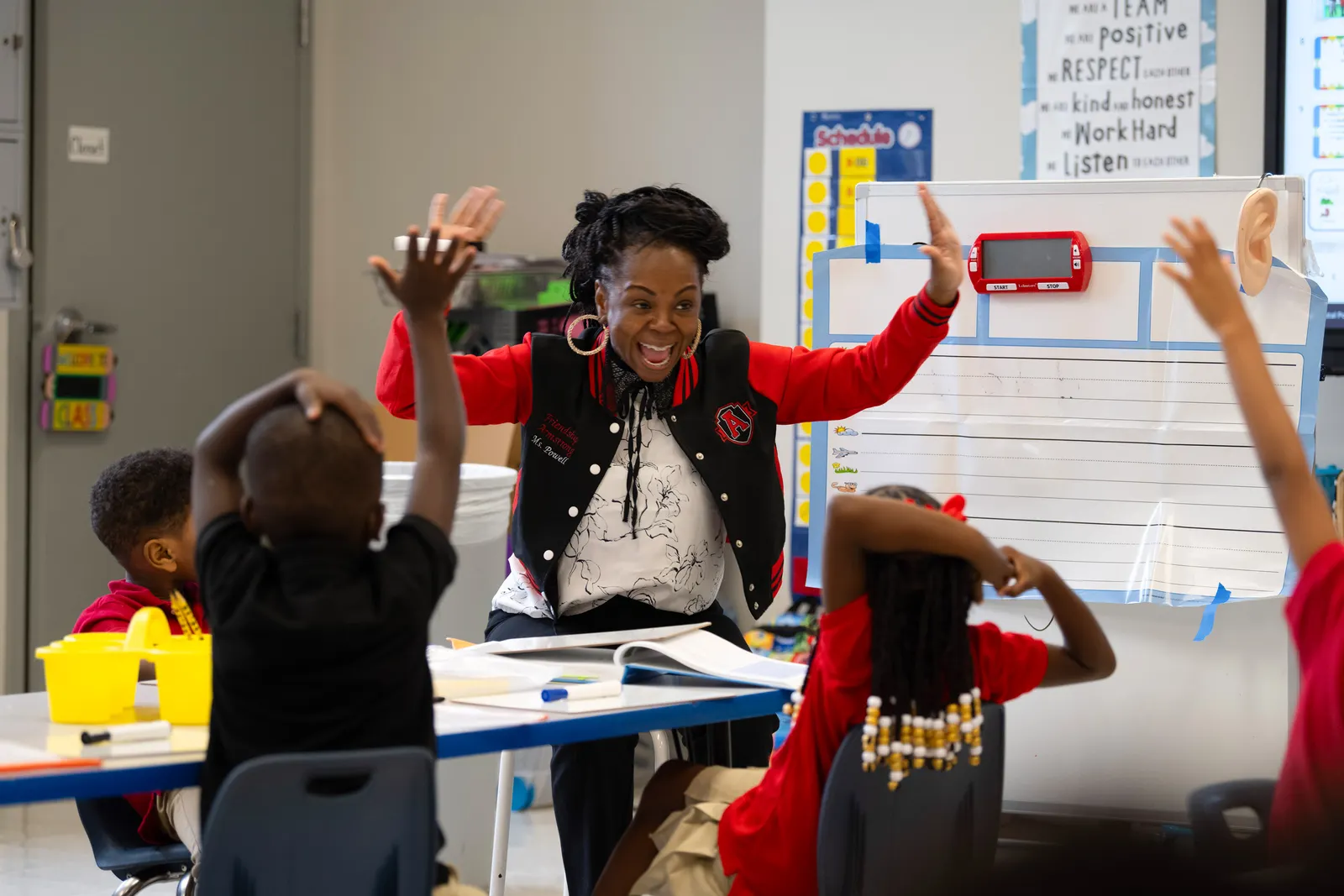 An adult is sitting at a table across from several young students whose backs are to the camera. The adult is smiling and has their arms in the air.