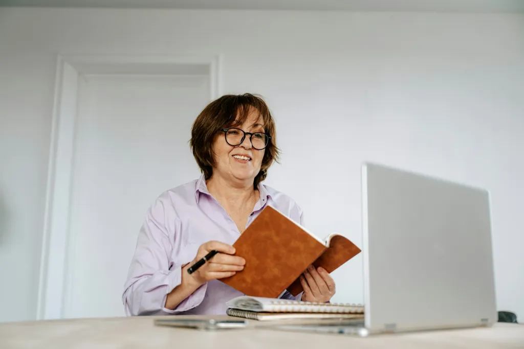 A middle-aged woman wearing a lilac button down top holds a notebook and pen. She's smiling at the open laptop on her desk as if she's on a video call.