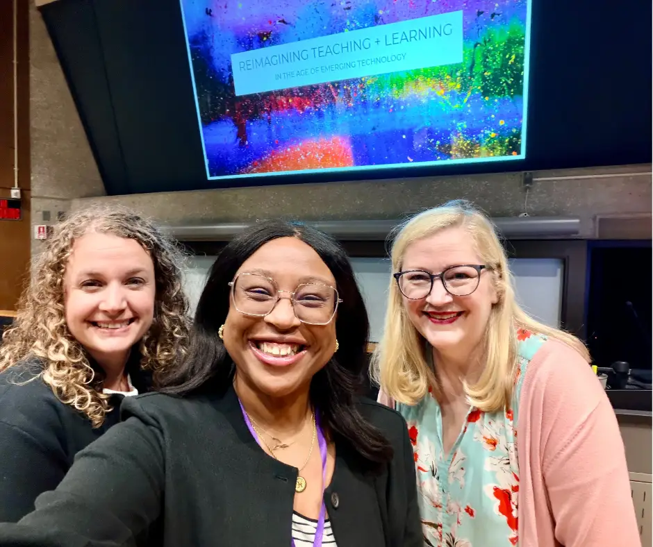 Three women stand at the front of a large lecture hall in front of a colorful presentation slide 