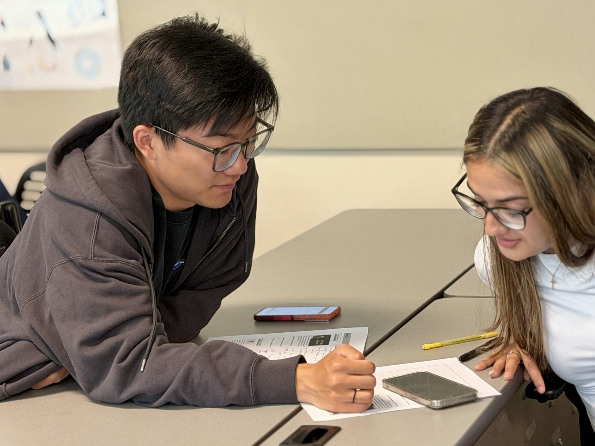 two students wearing glasses sit at at table looking at a paper
