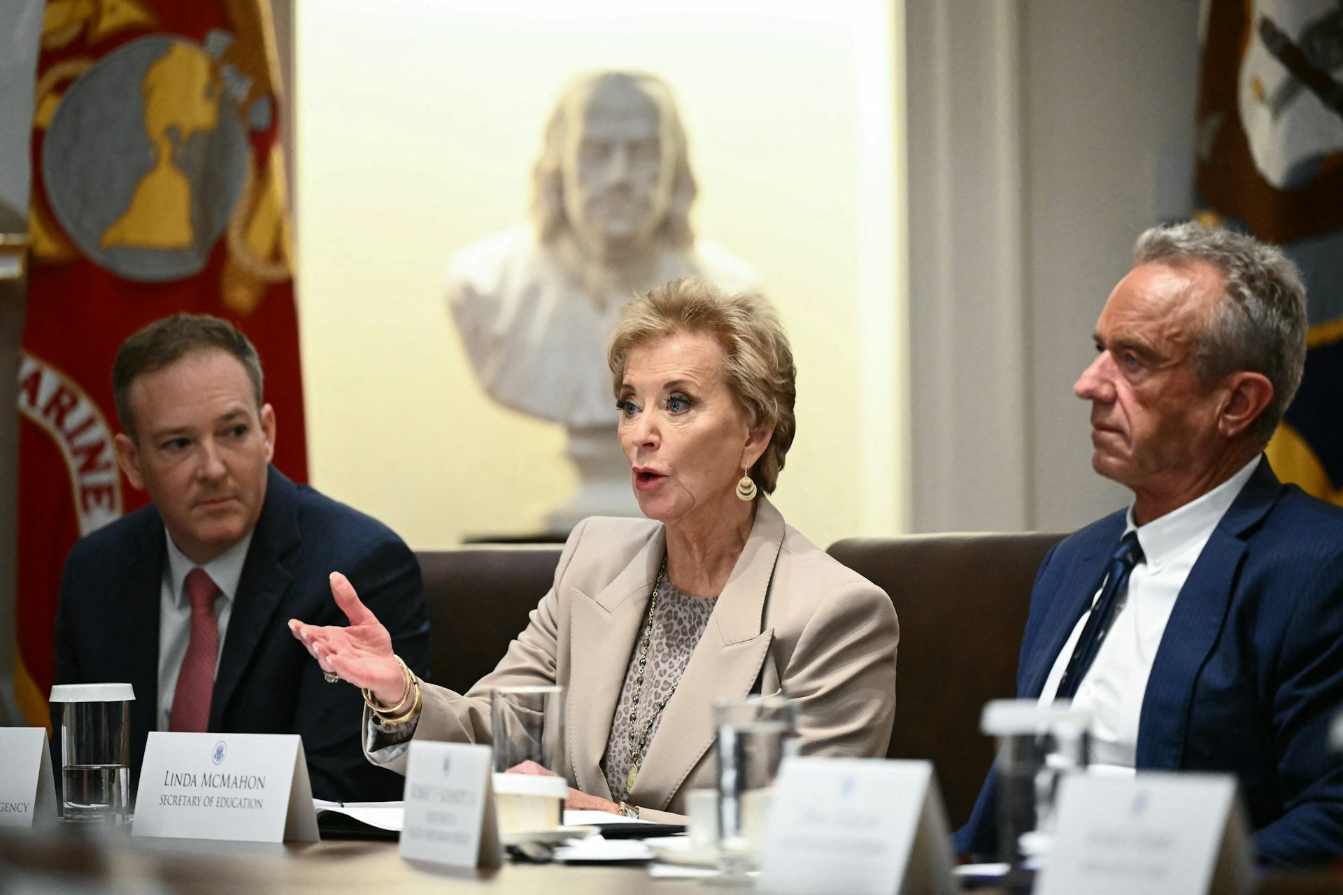 An older white woman wears a cream suit and sits at a table, with two men on either side of her.