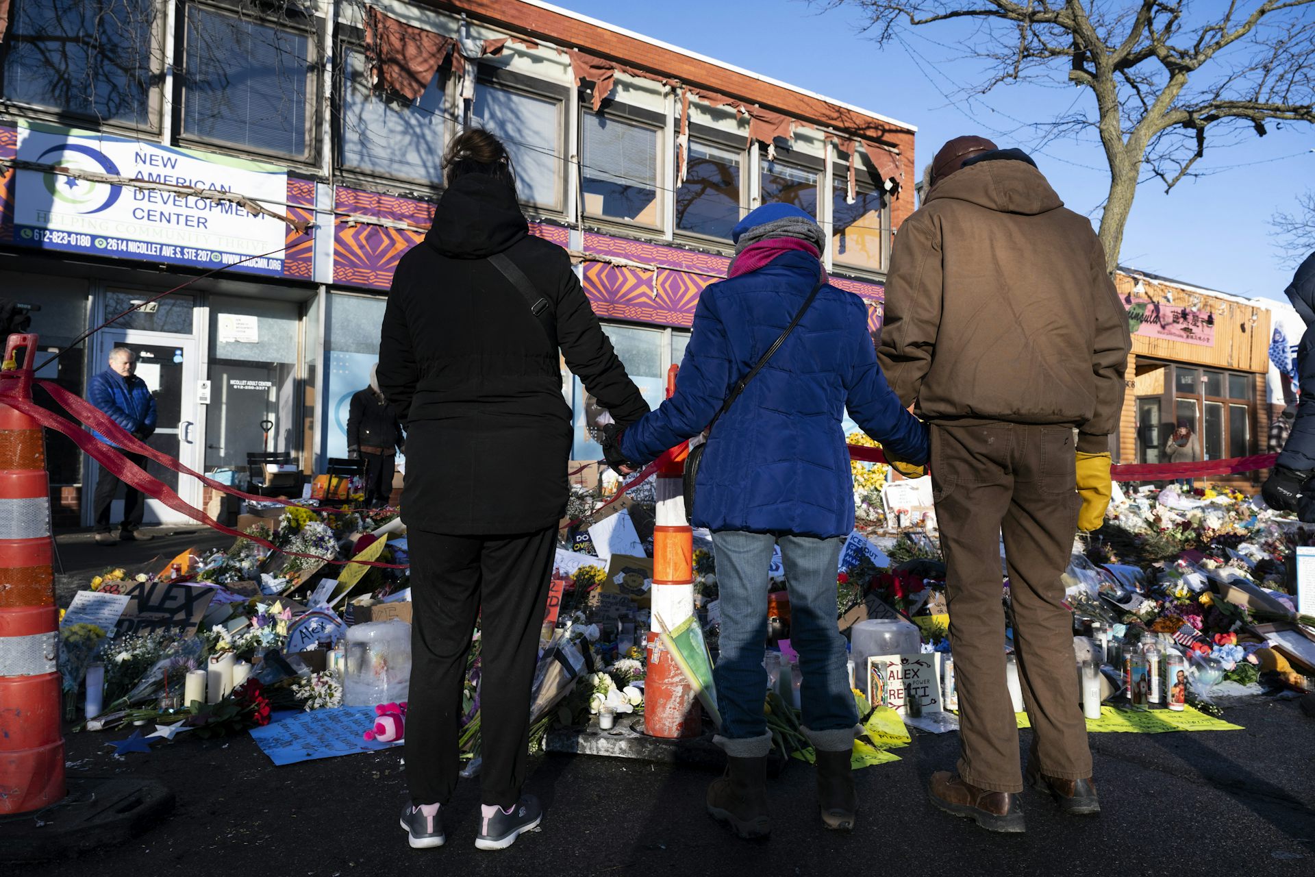 Three people, as seen from behind, stand and hold hands in front of a large memorial on a street that has flowers.