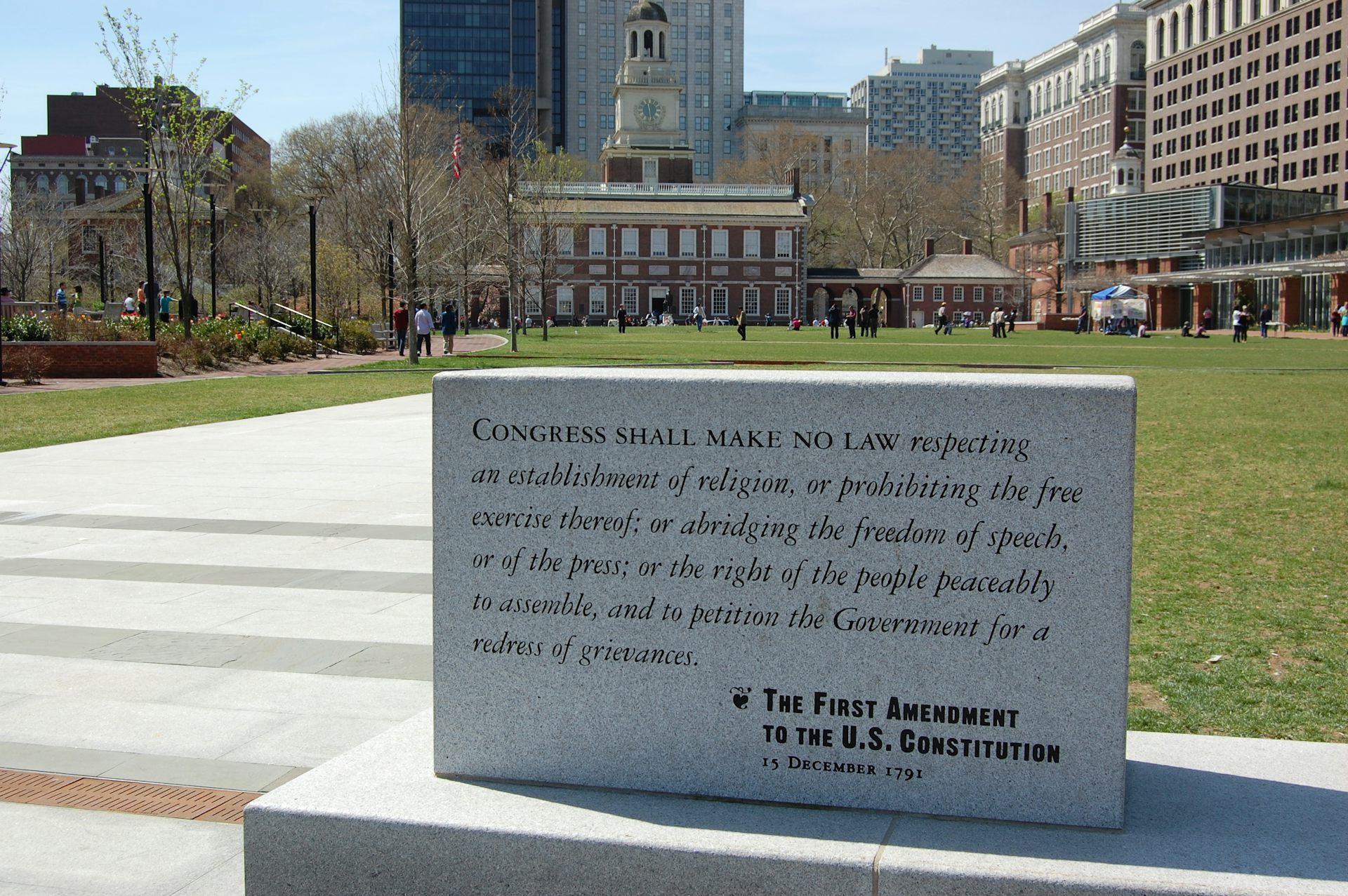 A large gray stone plaque shows the First Amendment in front of a green grassy field and buildings in the distance.