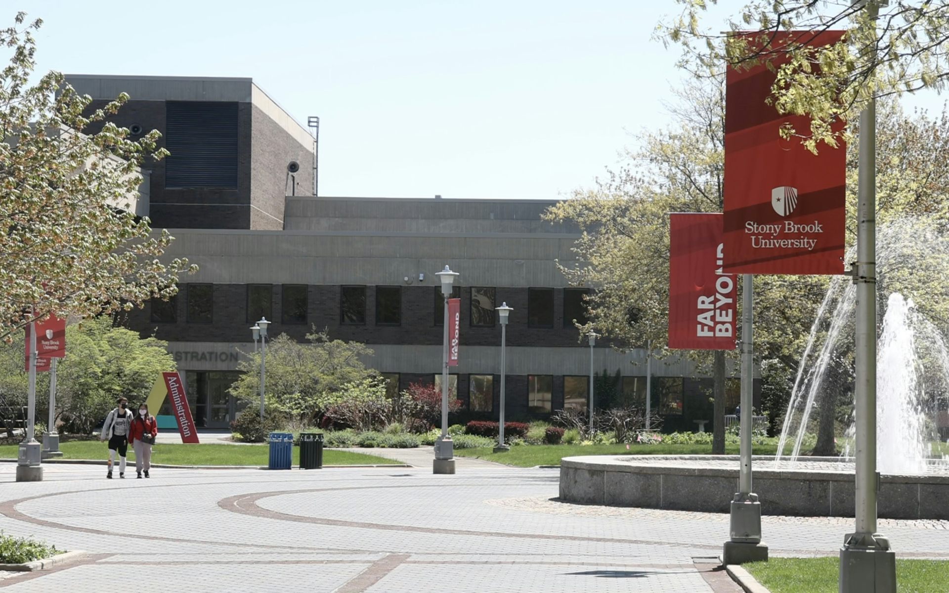A gray building is seen with red signs hanging nearby that say 'Stony Brook University.'