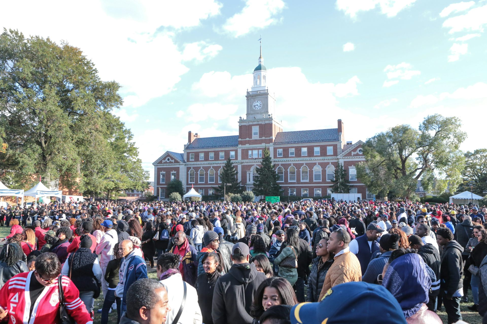 A large crowd is seen on a field in front of a red brick building with a tall clock tower.
