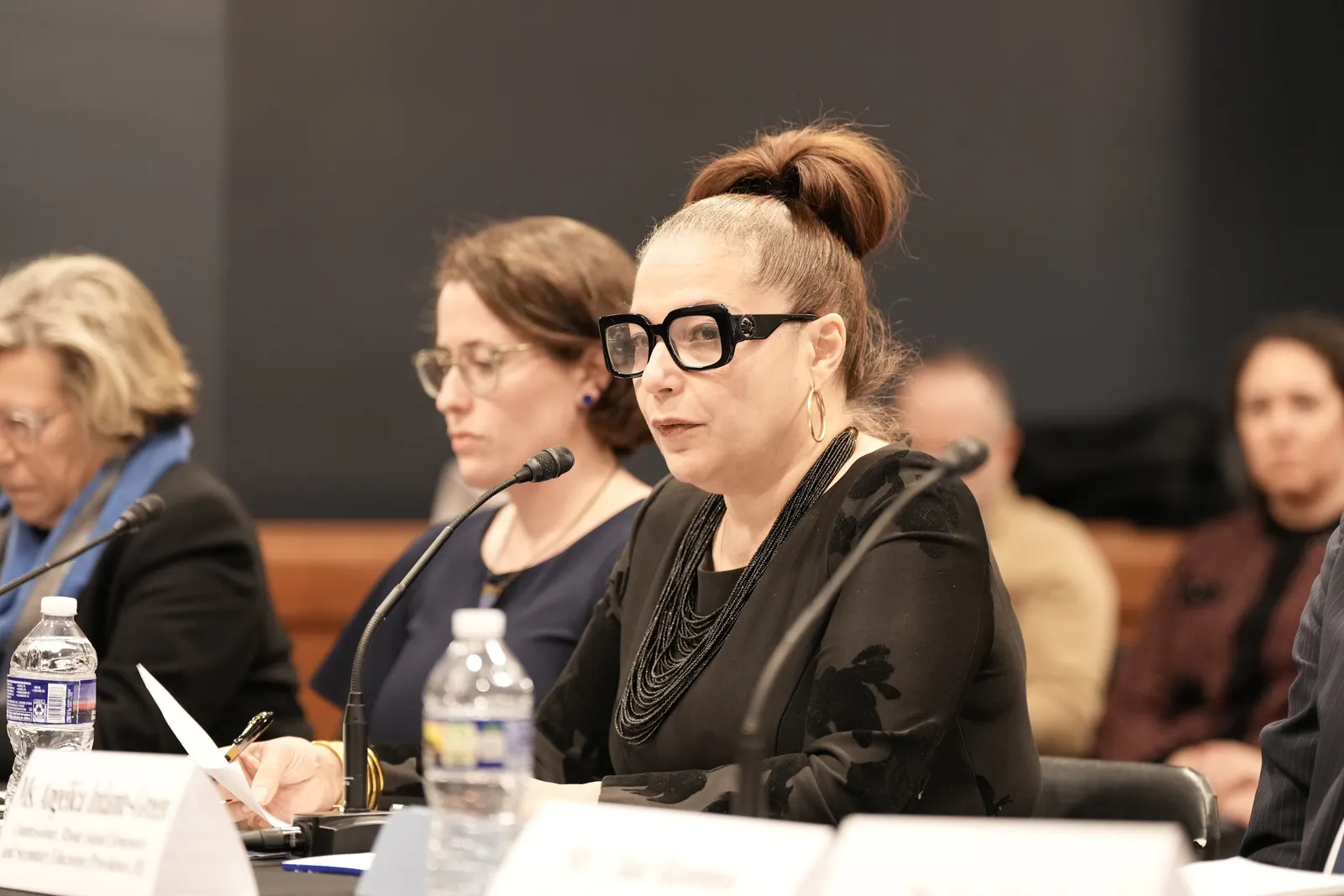 An adult sits at a table with a name tag and microphone in front of them.