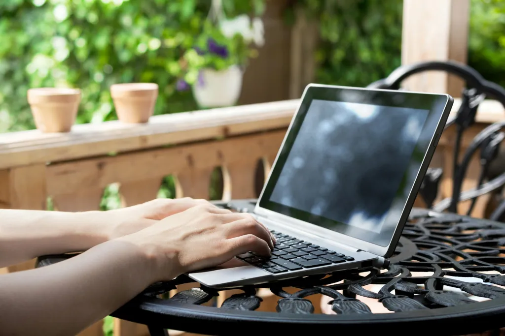Hands typing on an open laptop on an iron-wrought table on a balcony. It's a sunny day with green leaves and trees all around.