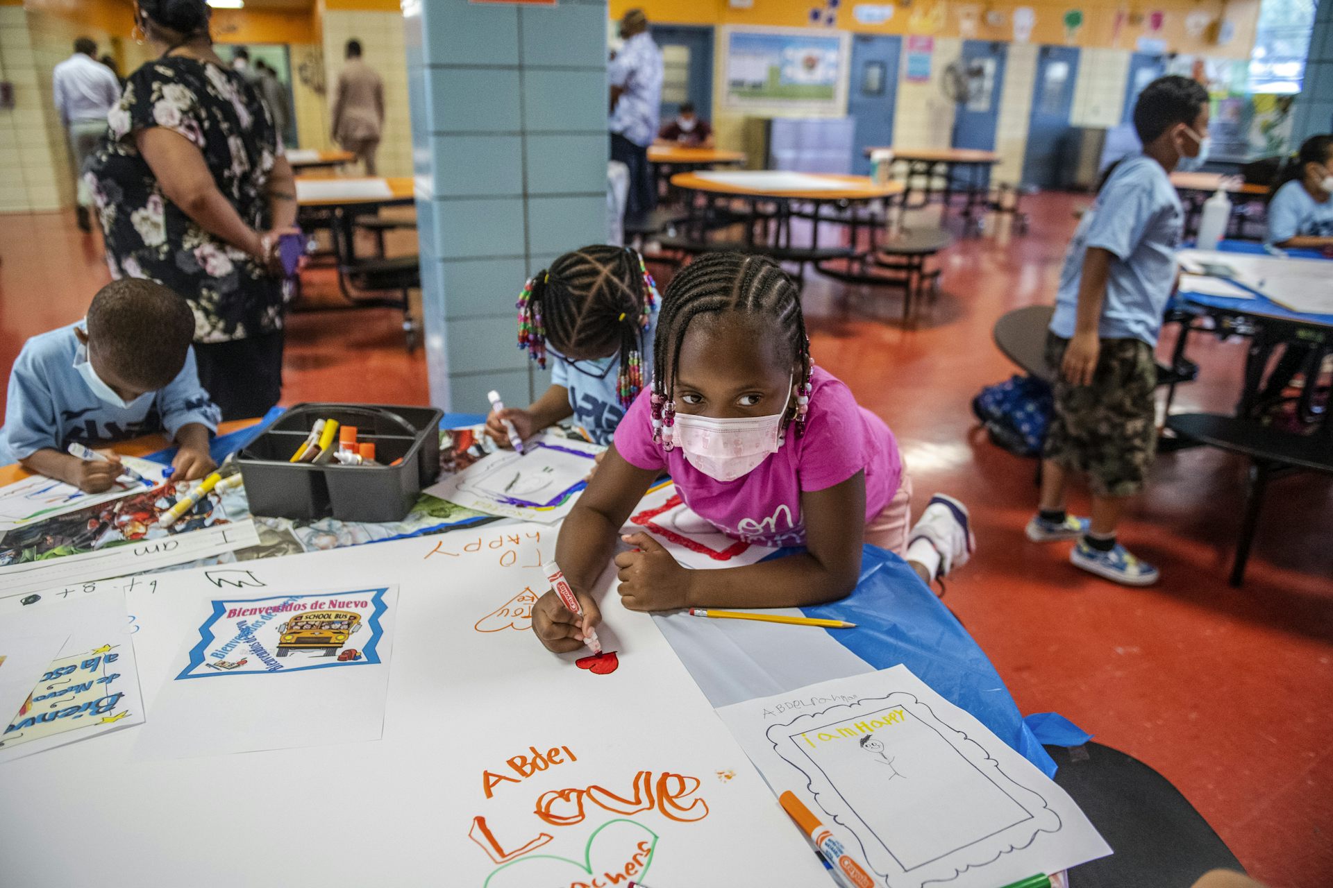 A group of young children wearing face masks sit at a table and color on white paper.