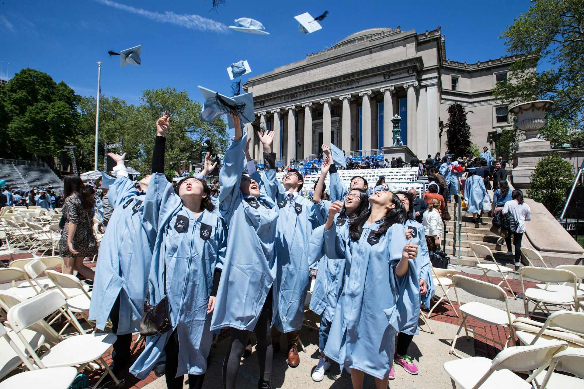 A group of young people wear light blue graduation robes and throw their caps into the air. They stand outside of a large building with columns.