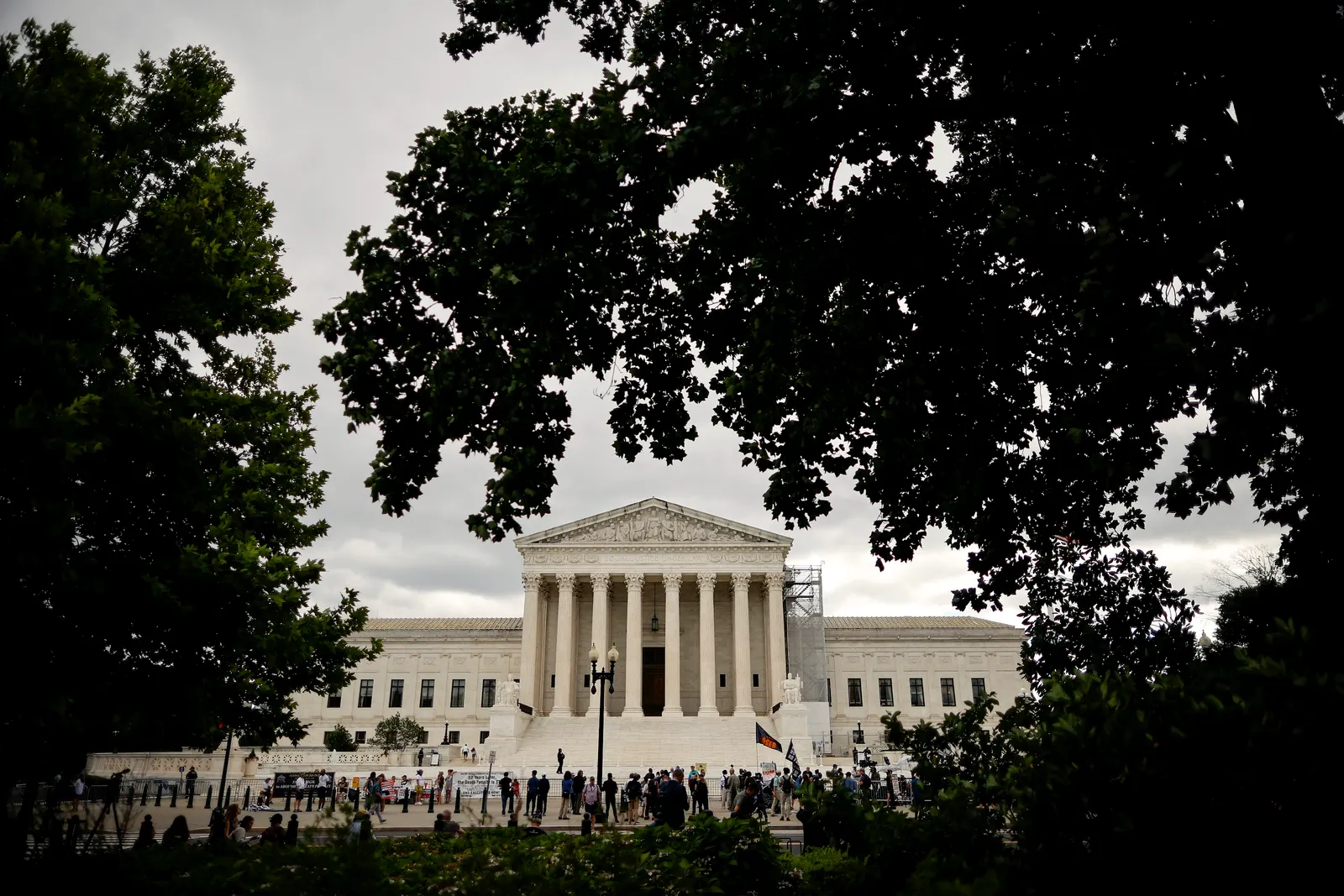 The facade of a large white building framed by trees is seen. People are gathered in front of the building.