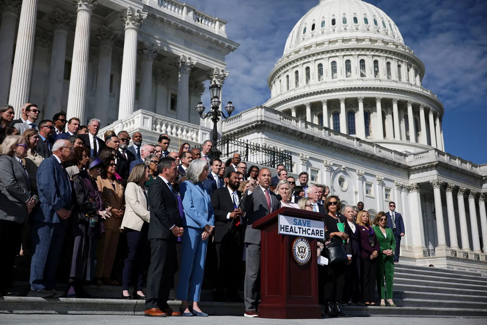 A large group of House representatives stand on the steps of the U.S. Capitol behind a podium that says "Save Healthcare."