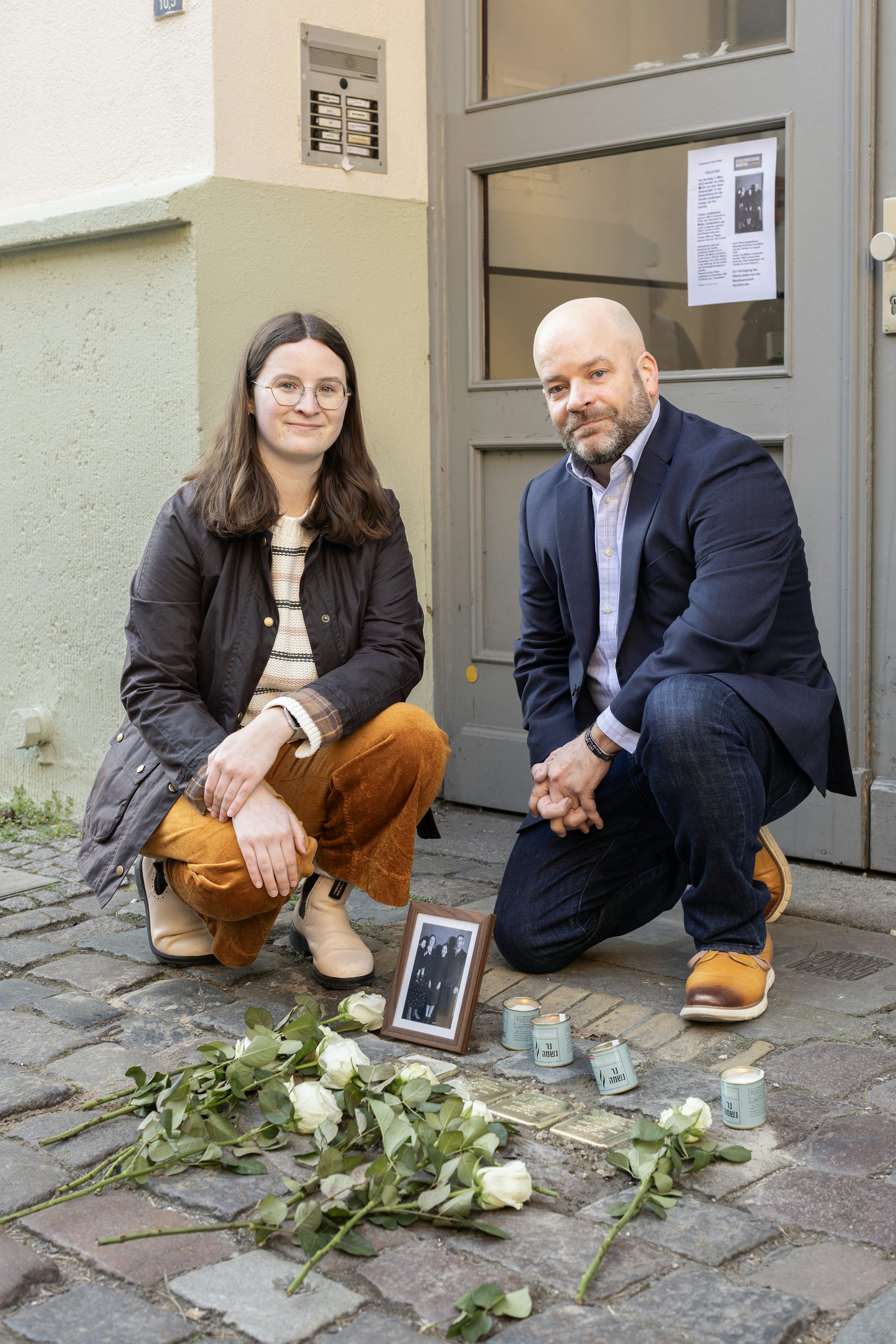 A young woman and a man in a blue suit kneel by a small memorial installed on a sidewalk of paving stones.