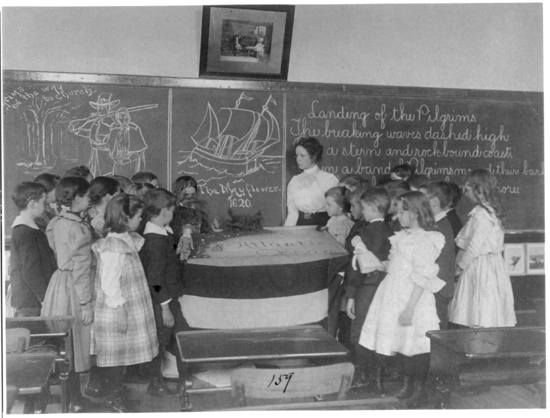 A black and white photo shows children dressed formally and standing around a table and a chalkboard with a woman standing near them.