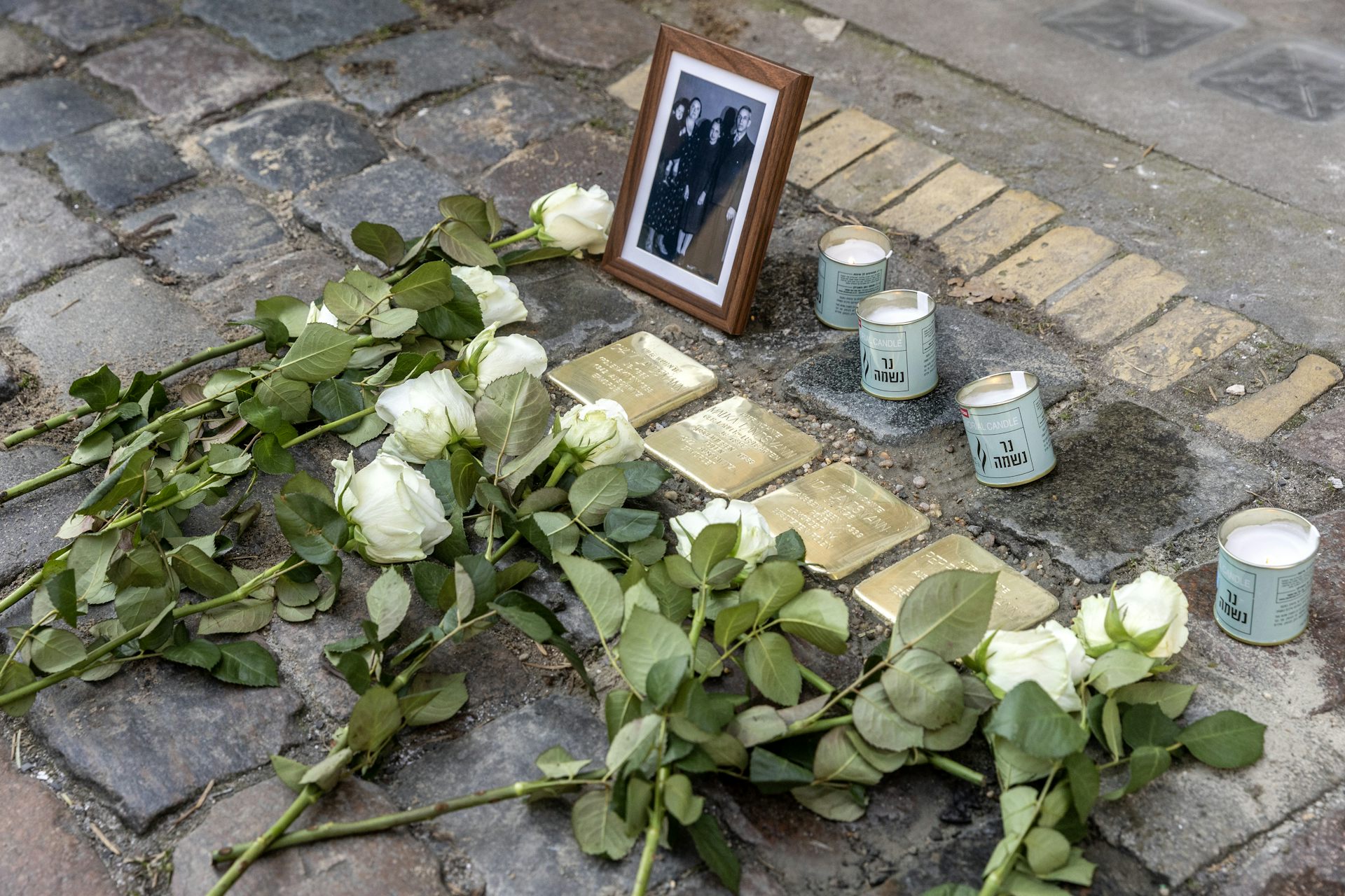 White roses placed on a sidewalk with four inlaid, bronze memorial stones, next to four candles and a framed family photo.