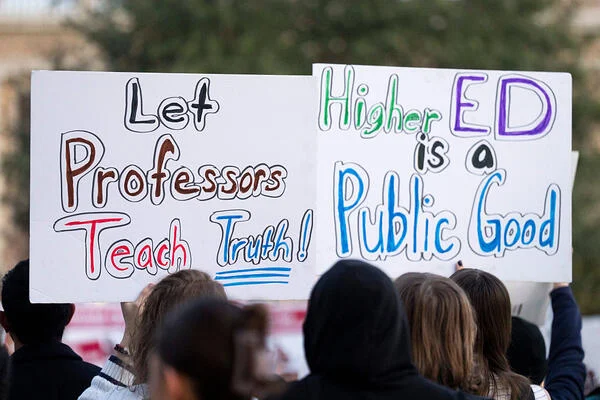 Protesters hold two signs on white poster board that say Let Professors Teach Truth and Higher Ed is a Public Good